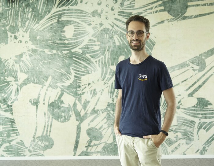 Mezzo busto di Alex Casalboni. Il ragazzo indossa una t-shirt blu con il logo AWS a destra, e un paio di occhiali. Sorride verso la camera. Sullo sfondo, una carta da parati beige e verde.