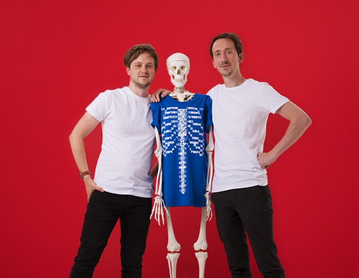 Ed Barton e Ben Kidd, fondatori di Curiscope. I due ragazzi, in t-shirt bianca e pantaloni nero, abbracciano uno scheletro con una t-shirt blu. Sfondo rosso.
