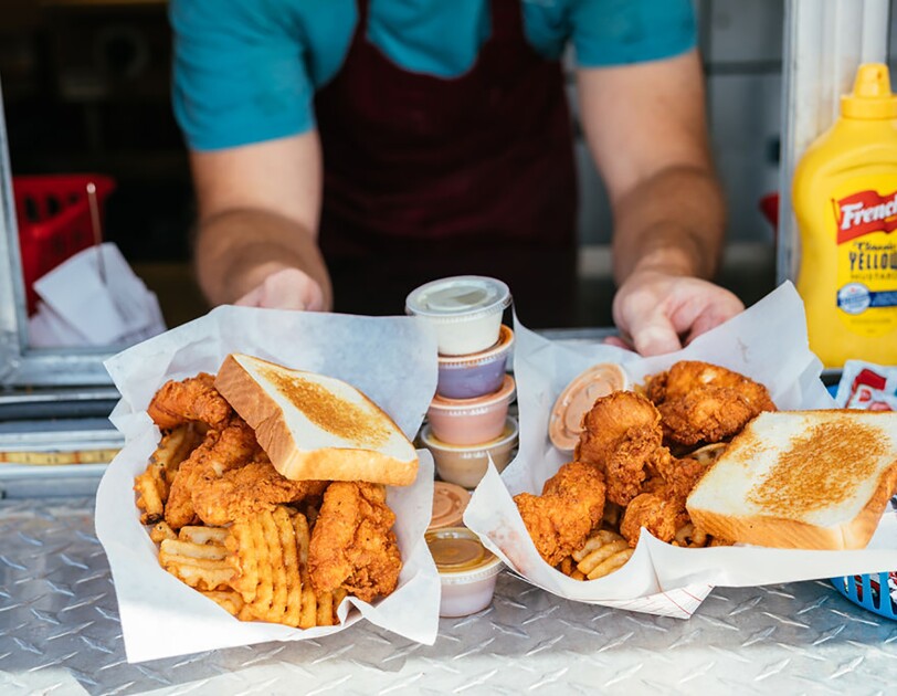Baskets of chicken fingers, waffle fries and Texas toast are passed through the window of a food truck. A stack of condiments and sauces balances between the two baskets of food.