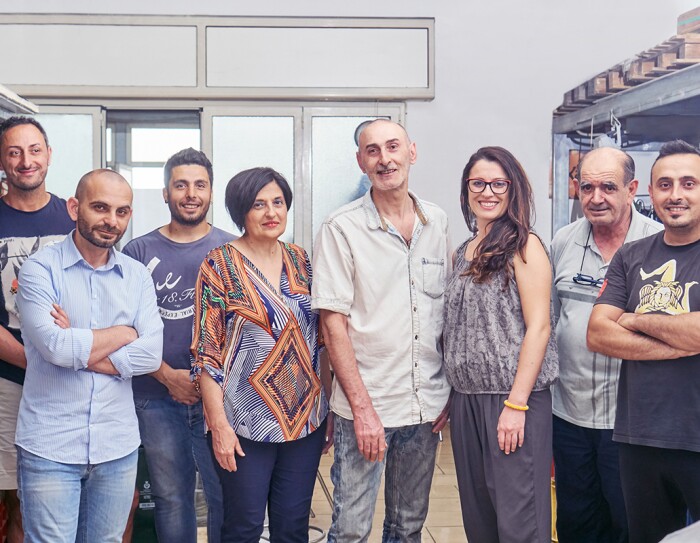 Foto del team di Kamira, formato da 8 persone in piedi, 6 uomini e 2 donne, tutti sorridenti, inclusi il fondatore Nino Santoro e la sua famiglia.