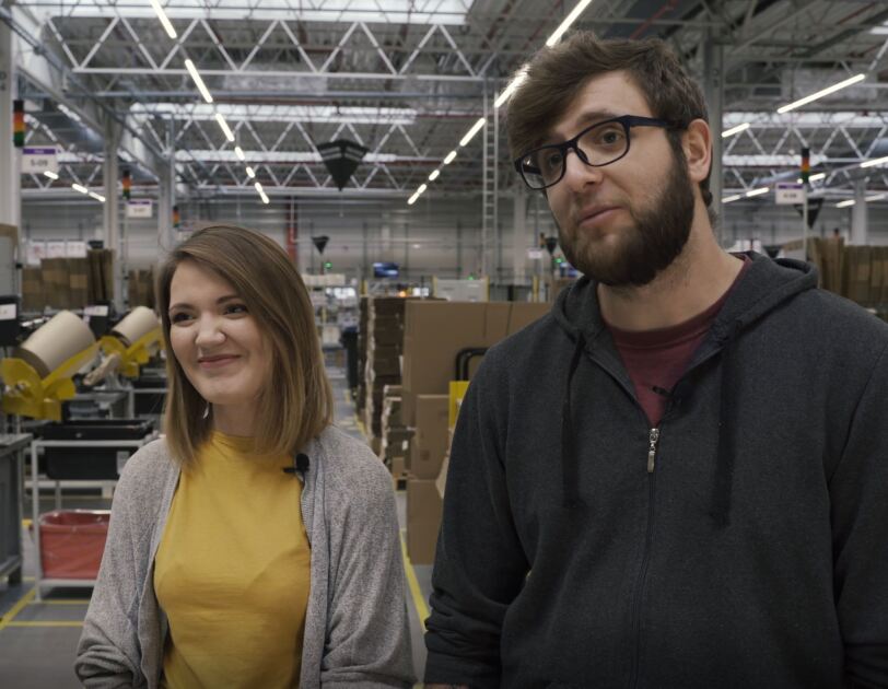 Une femme se tient dans un centre de distribution Amazon, avec un sourire sur son visage.  &Agrave; sa gauche, un homme se l&egrave;ve.