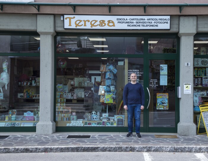 Andrea Locatelli posa di fronte all'ingresso del negozio di Teresa. L'insegna dice "Teresa - edicola, cartoleria, articoli regalo, profumeria, servizio fax, fotocopie, ricariche telefoniche."