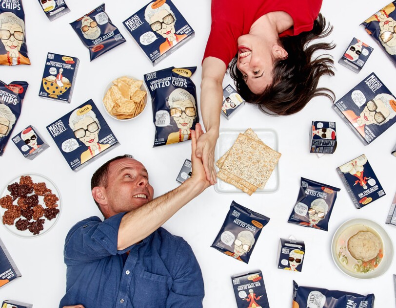 A woman lies on her back, with her right hand extended above her head as she gives a man, also lying on the ground, a high five. Around them are packages of matzo and other products.