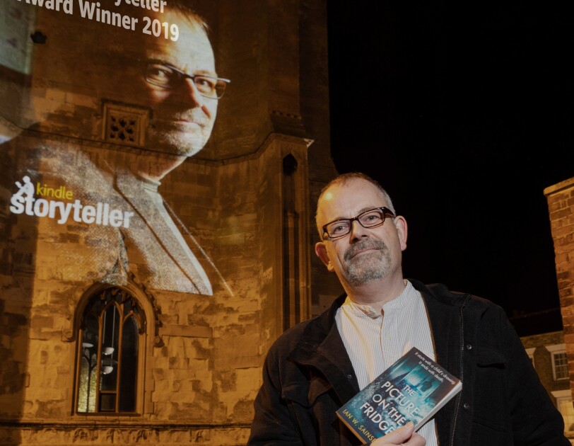 Ian W. Sainsbury holding his book in front of a Storyteller projection of his head shot congratulating him on his award.