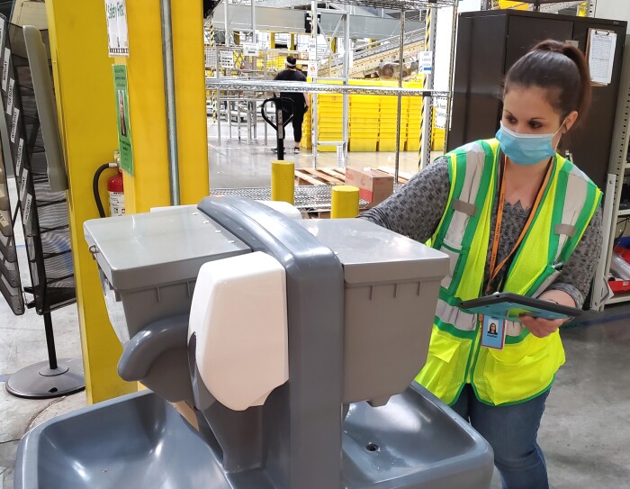 A woman wearing a safety vest and mask inspects a hand-washing station.