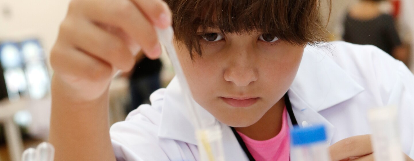 Un chica de unos 11 años, con el pelo recogido y bata blanca, está colocando en una probeta un líquido. Tiene la mirada fija a la probeta y en la mano tiene una pipeta. Es un primer plano de la chica, El fondo difuminado.