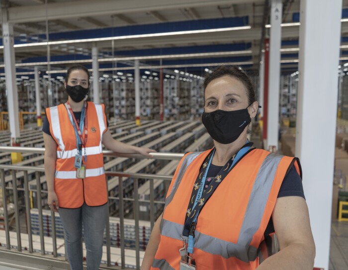 Marta Torrejón Serrano y María Jesús Serrano. Madre e hija posan en un balcón del centro logístico de Illescas que se ve de fondo. Están apoyadas a la banadilla manteniendo la distancia de seguridad. Las dos van con manga corta, mascarilla corporativa y chaleco naranja. Llevan el pelo recogido con una coleta.