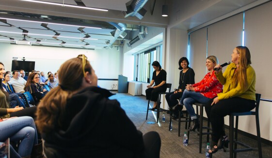 Four women are seated at the front of a conference room, leading a panel on leadership. Each is seated on a raised bar-height stool. In front of them, Dozens of Amazonians face them, as they speak.