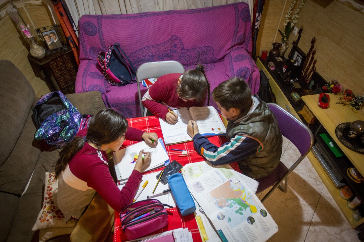 Tres niños, dos niñas y un niños, trabajando en una mesa situada en el salón de una casa. Dos de ellos están sentados enuna silla y la tercera está sentada en el sofà. Dos de ellos trabajan sobre el mismo libro. Hay dos sofás y en la mesa hay libros, lápices y rotuladores. Los tres van muy abrigados y tienen las mochilas tiradas por el sofá.