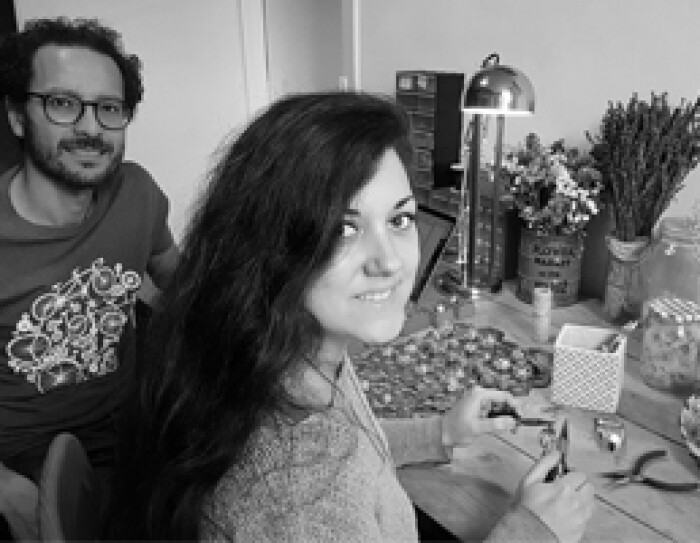 Les deux fondateurs de Trois Fenêtres se tiennent devant une table où est posé du matériel de création artisanale ainsi qu'une lampe et des bouquets de fleurs. C'est une photo noir et blanc.