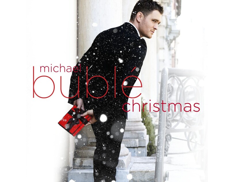 Michael Buble stands outside wearing a tuxedo, facing away, but seeming to be glancing over his shoulder. he holds a small, wrapped gift in red paper with black bow. Snow is falling in the image, and text saying "Michael Buble Christmas" is written over the top of the image.