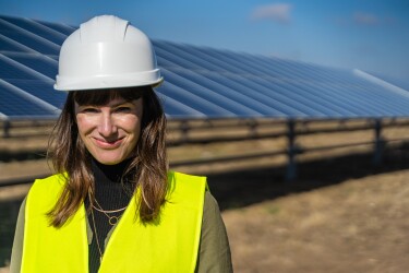 Denise Hugo, jefa de proyectos de Energías Renovables de Amazon. Ella está delante de las placas solares. Va vestida con casco blanco y chaleco amarillo. Tiene el pelo castaño y le llega más allá de los hombros. LLeva tres collares de oro que están por encima de un jersey de cuello alto marrón.