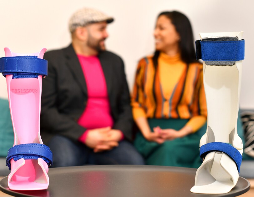 In the forefront, two ankle orthoses for children produced by Andiamo, a smaller one in bright pink plastic and a bigger one in white plastic with blue straps. In the background, Samiya and Naveed Parvez smile at each other.