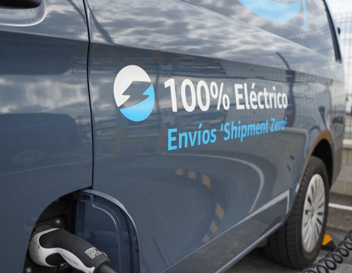 Una Mercedes Benz-Vans en un aparcamiento. Se ve el logo Prime de Amazon y el texto en la puerta 100% Eléctrico Envíos 'Shipment Zero'. En primer plano se ve un cargados de batería que está subministrando electricidad a la furgoneta de al lado que solo se ve un poco su parte trasera.
