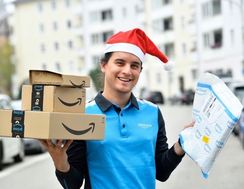 Ein junger Mann in Amazon Zustellkleidung und roter Zipfelmütze hält eine Reihe von Paketen in der Hand