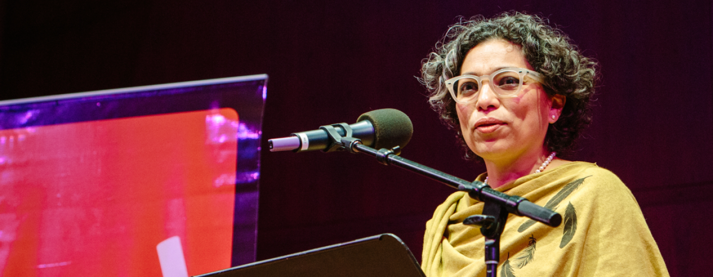 A woman standing at a lecture, in front of a portable podium and microphone. She appears to be speaking to a group, on a stage. In the background, Seattle Arts & Lectures logo is visible.