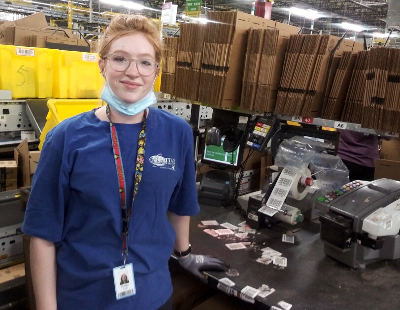 A woman stands at a workstation stocked with cardboard boxes and labeling machines. She has a protective mask on, ready to cover her mouth and nose.