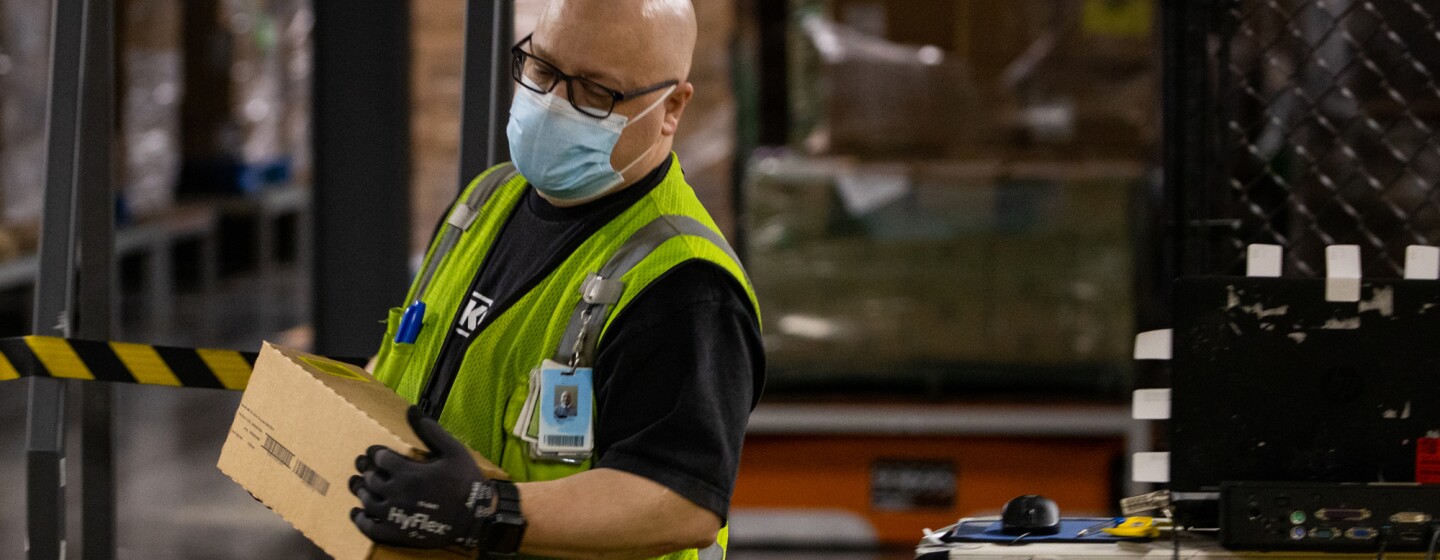 A man wearing gloves, a mask, and a safety vest carries a small box.