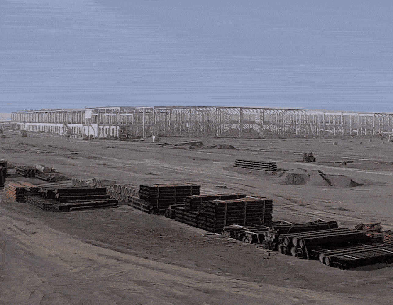 El centro logístico de Illescas en construcción. Dos trabajadores con cascos, blanco y azul, y chalecos amarillos aparecen apoyados en una mesa trabajando en un plano. Uno de ellos lleva un guante blanco y señala el mapa. De fondo se ve una grúa trabajando.
