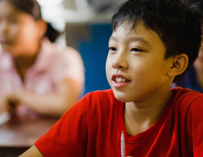 Ein asiatischer Junge im roten T-Shirt sitzt in der Schulbank. Im Hintergrund sieht man weitere Schulkinder, die am Unterricht teilnehmen.