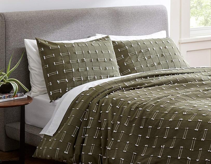 A bed in a home setting - the bed has a gray padded headboard, white sheets, and olive green shams and duvet. Next to the bed is a side table with two books and a plant.