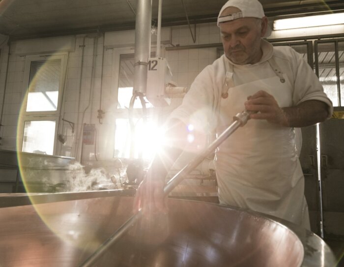 Immagine di un uomo mentre lavora il formaggio nel laboratorio della Latteria Due Madonne. L'uomo regge lo strumento chiamato 'spino' e con questo gira la cagliata dentro un grosso pentolone. L'uomo è vestito di bianco e indossa un grembiule e un cappello. La scena è ripresa di giorno.