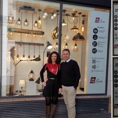 Ein Mann und eine Frau stehen vor einem Schaufenster, in dem viele unterschiedliche Lampen zu sehen sind. Beide lächeln in die Kamera.