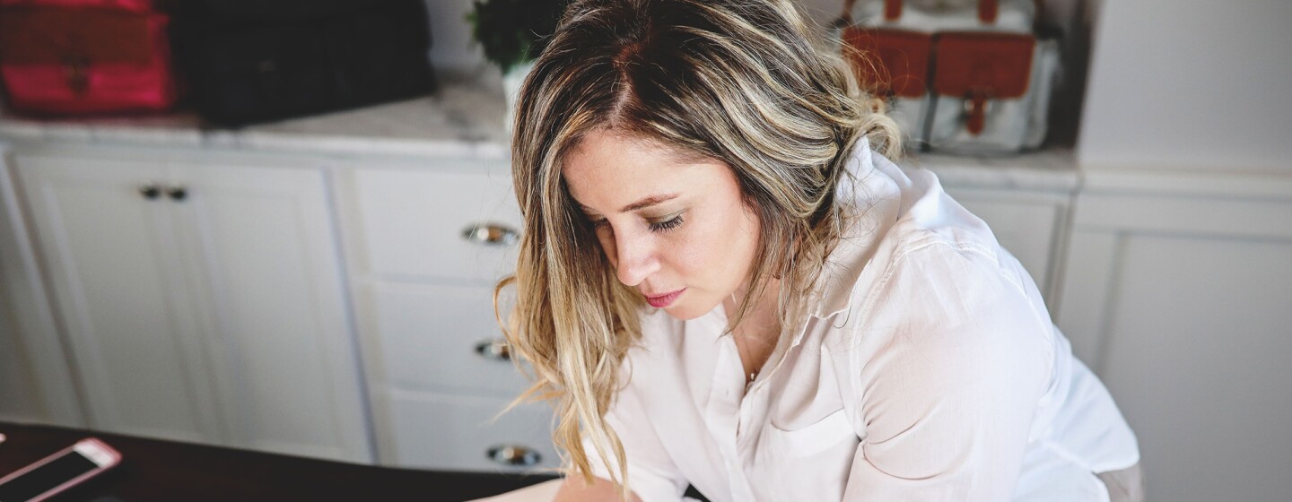 A woman wearing a white button down and jeans, leans over a book of fabric swatches. To her right is a computer and mobile phone. Behind her is built-in cabinetry.
