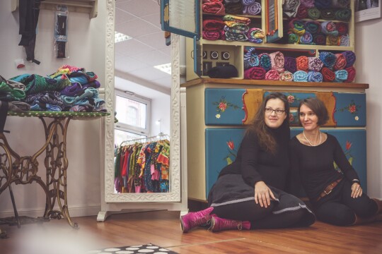 Die beiden Gründerinnen von Mardame Jordan sitzen in ihrem Geschäft. Im Hintergrund sieht man viele bunte Babykleidung.