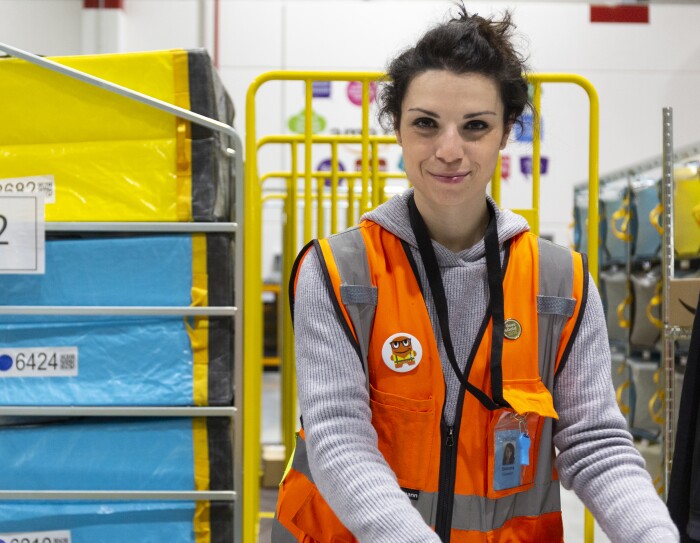 Simona Calzolari, operatrice di magazzino, inquadrata nel deposito di smistamento di Brandizzo. Indossa un giubbotto arancione ed è circondata da carrelli gialli e pacchi celesti e gialli.