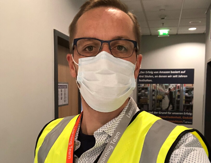 Ein Mann mit blonden Haaren, Brille und Sicherheitsweste vor einer Packstation im Logistikzentrum