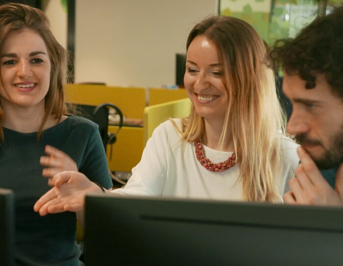 Dos mujeres y un hombre, todos de unos treinta años. Las dos mujeres están sonriendo mirando una pantalla de ordenador y el hombre, con la mano apoyada en su barbilla, observa la pantalla.