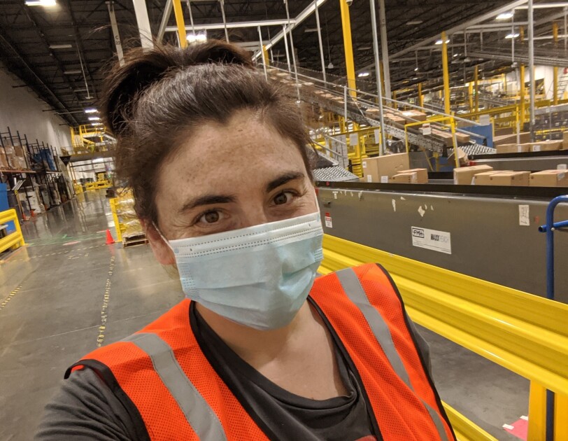 Sarah Freeman, an Amazon associate, is smiling for a selfie inside the Amazon fulfillment center where she works. She is wearing a mask and other Amazon safety gear like an orange vest.