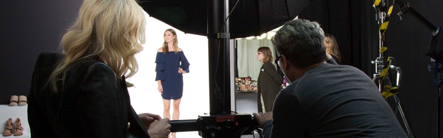 Two individuals, a male photographer and a woman in a leopard-print dress and dark jacket draped over her shoulders, photograph a model wearing a blue dress during an Amazon Fashion shoot.