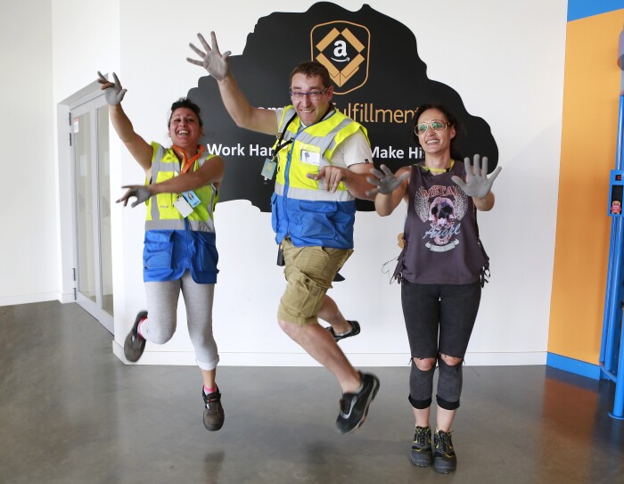 Patricia Alonso, Maribel Salcedo y Marius Dumitrescu aparecen en la entrada del centro logístico saltando y mostrando las palmas de sus manos pintadas de color gris. Están sonriendo mirando a cámara. Marius se enceuntra en medio.