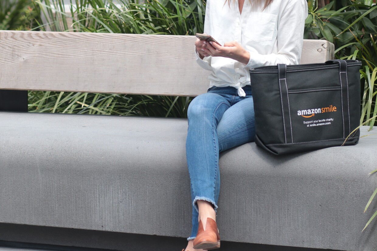 A woman on a bench holds a smartphone. She is seated next to a tote bag with the AmazonSmile logo and the words "Support your favorite charity at smile.amazon.com."
