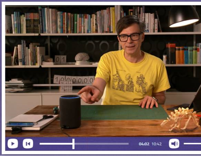 Ein Mann in einem gelben Tshirt sitzt an einem Schreibtisch und zeigt mit dem Finger auf ein Alexa Echo.