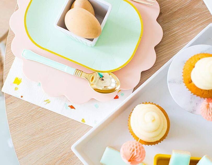 A white melamine tray sits on a light wood table. On the tray sits a small yellow dish of ice shortbread cookies, a small cupcake, and a small marble-looking cupcake stand. To the left of the tray, a small, scalloped plate sits under a gold-rimmed aqua plate, which holds a small striped white dish.