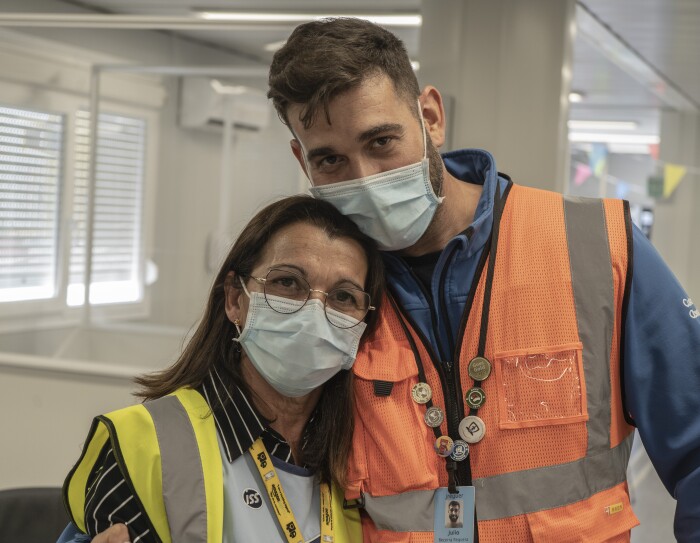 María José Requera y Julio Becerra Requera. Situados en las oficinas de la estación logística de Amazon en Málaga. La madre va con un chaleco amarillo, camisa negra con rayas blancas. Lleva gafas, mascarilla quirúrgica y el pelo suelo por el hombro. Su hijo le pasa la cabeza, lleva mascarilla quirúrgica, el pelo corto, un forro polar azul y un chaleco naranja.
