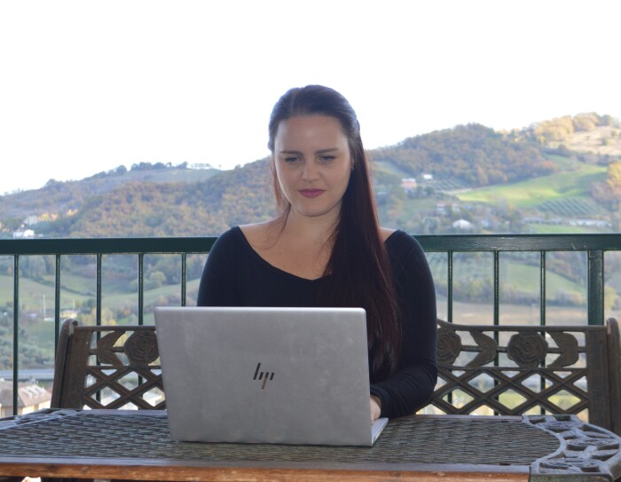 Una donna è seduta ad un tavolo su un terrazzo e ha di fronte a sè un PC. Alle sue spalle delle colline.