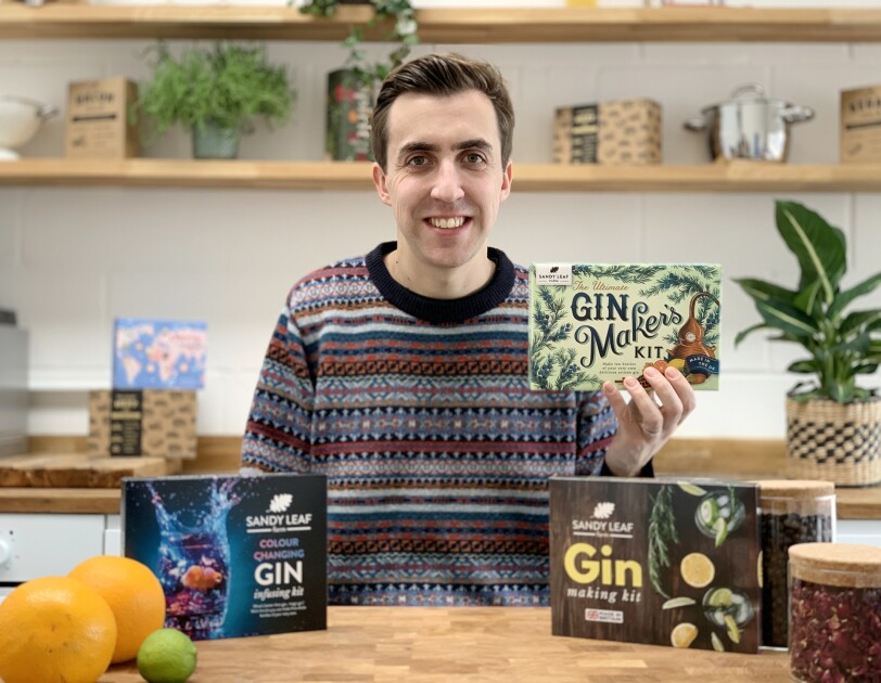Scott MacDonald from Sandy Leaf Farm at a table with the gin making kits in front of him. He is also holding a box containing the products.