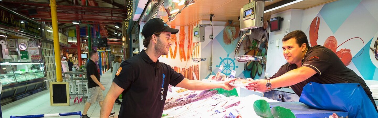 Mercado de La Paz de Madrid. Trabajador de Prime Now recoge en una pescadería el paquete de un cliente. El pescadero, con un delantal azul, tiene que incorporarse para entregarle el pedido. El trabajador de Prime Now va con un carro de la compra que contiene más pedidos.