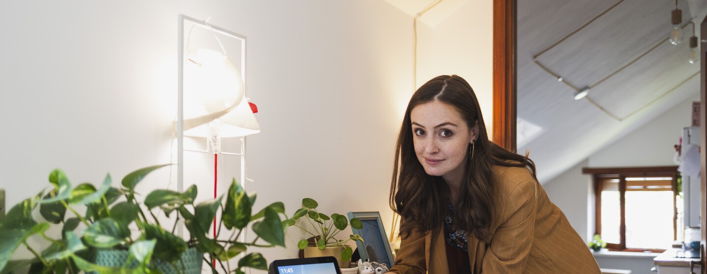 Elisa Reale, al centro della foto, è chinata verso un mobile. Guarda in camera, vicino alla mano destra ha un dispositivo Echo Show con lo schermo acceso. In primo piano, posata sullo stesso mobile, si vede una pianta in un vaso verde. Elisa indossa una giacca beige a righe bianche, ha i capelli lunghi e mossi. Sotto la giacca indossa una gonna a fiori colorata.