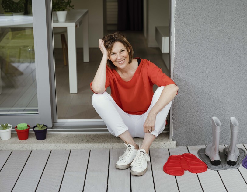 Eine Frau mit rotem T-shirt und weißer Hose sitzt in einem Türrahmen und lächelt in die Kamera.