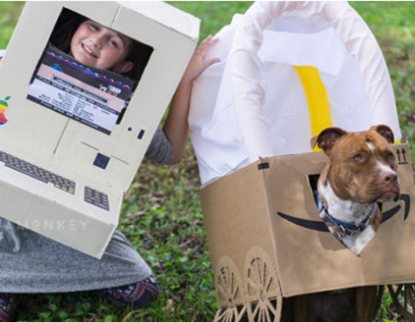 Halloween costume made out of Amazon boxes. On the left, a little girl wears a gray dress and a homemade apple computer from the 1980s. To her left is a dog in an Amazon box made to look like a covered wagon.