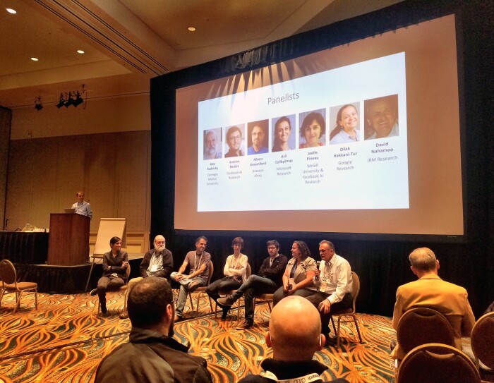 Scientists from academia and industry participate in a panel discussion at the recent NIPS Conference Conversational AI workshop. The panelists are seated in front of the audience, and their pictures are projected onto a screen.