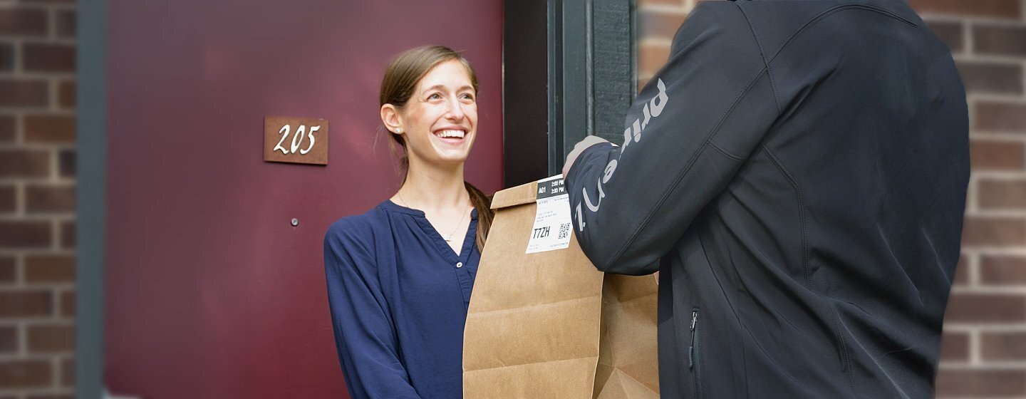 Repartidor de Prime Now con un chaqueta negra entrega una bolsa de papel de Amazon a una señora con una camisa azul que se encuentra en la puerta de su casa.