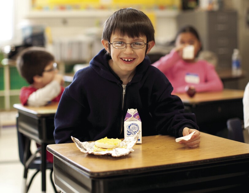 A boy sits at a school desk, smiling at the camera. On his desk are a carton of milk and a breakfast sandwich. Behind him, other students also eat and drink.