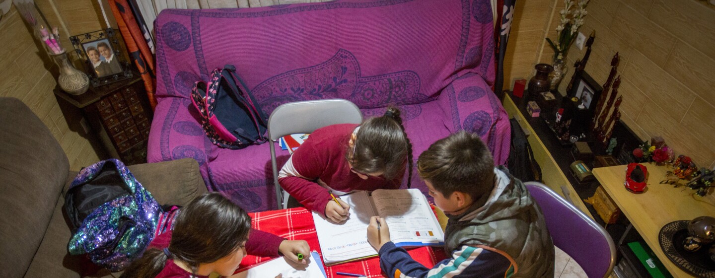 Tres niños, dos niñas y un niños, trabajando en una mesa situada en el salón de una casa. Dos de ellos están sentados enuna silla y la tercera está sentada en el sofà. Dos de ellos trabajan sobre el mismo libro. Hay dos sofás y en la mesa hay libros, lápices y rotuladores. Los tres van muy abrigados y tienen las mochilas tiradas por el sofá.