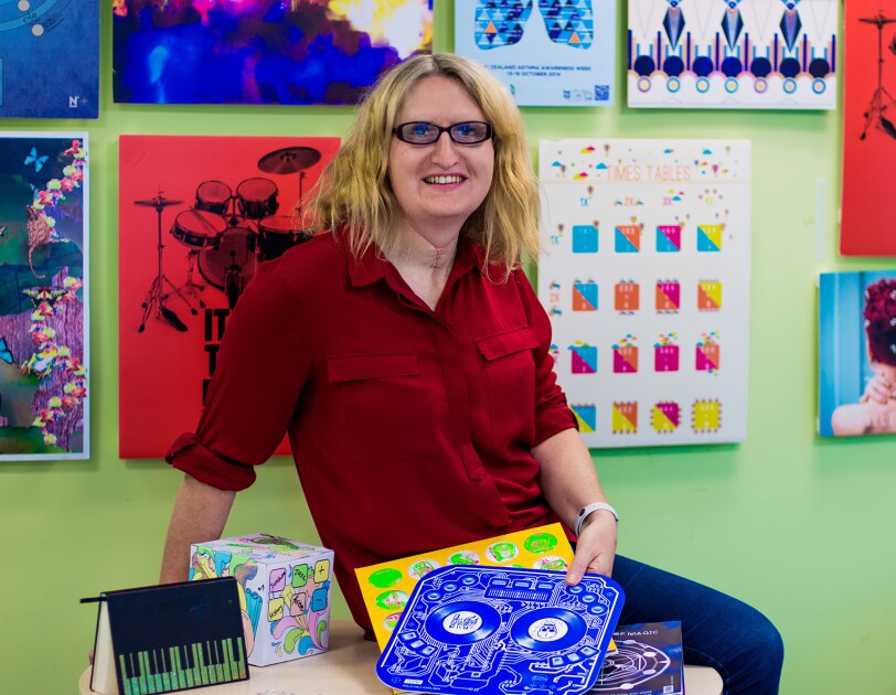 Eine Blonde Frau trägt ein rotes Hemd, Jeans und eine Brille. Sie sitzt auf einem Tisch und präsentiert ihre Produkte.